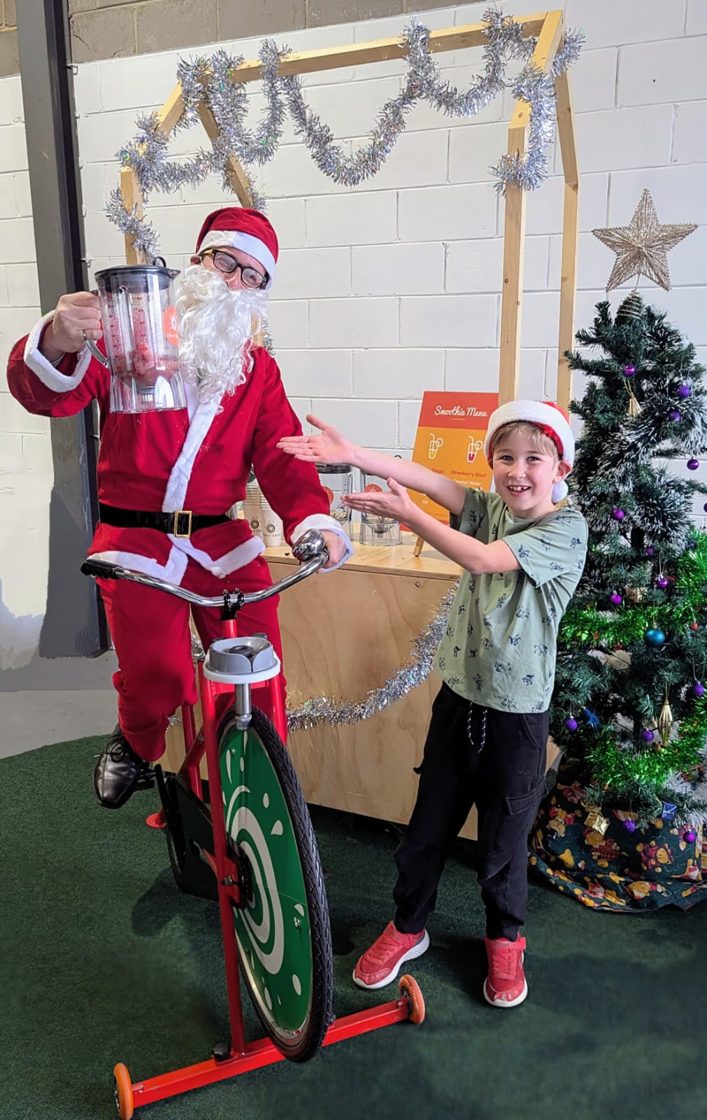 Santa on a smoothie bike holding a blender willed with a smoothie, wit ha child next to him having fun and wearing a Christmas hat