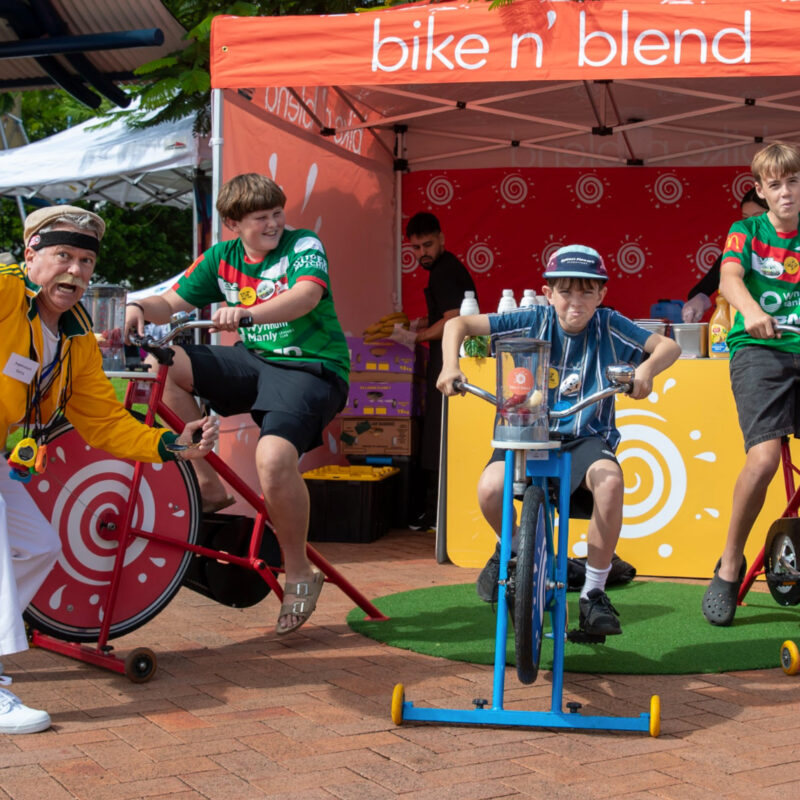 Kids having fun riding on the blender bikes in front of a colourful marquee, as an enthusiastic marching band leader cheers them on