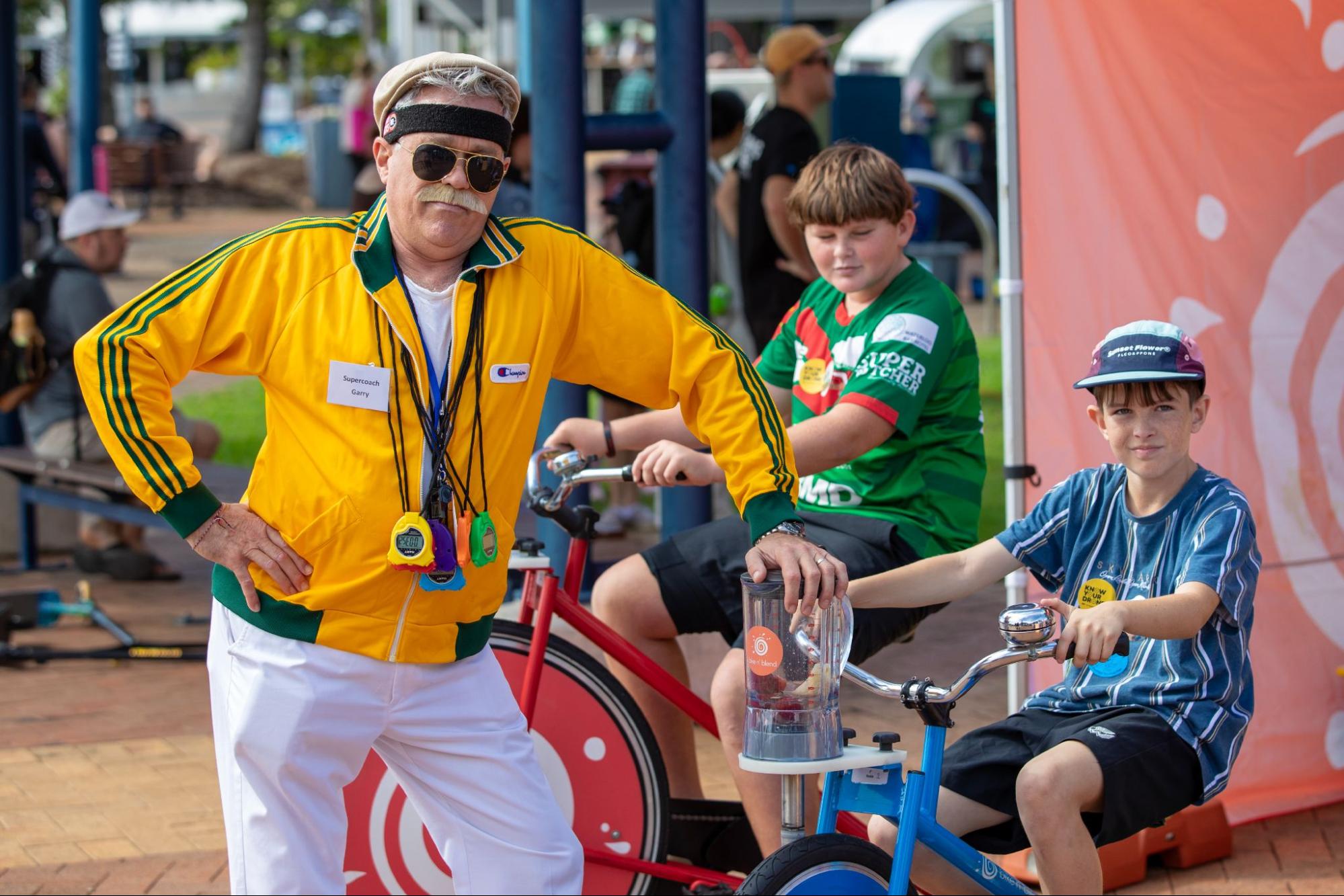 Colourful sports coach supporting two kids riding blender bikes, about to compete with one another